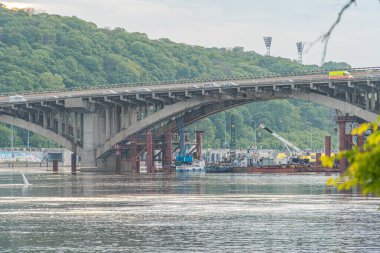 Gökyüzü bulutlu, Kiev 'deki Dnepr Nehri üzerindeki asma köprü, kırmızı destekli sanayi manzarası, çoklu kablolar, durgun su yansıması, yeşil ön plan santrali