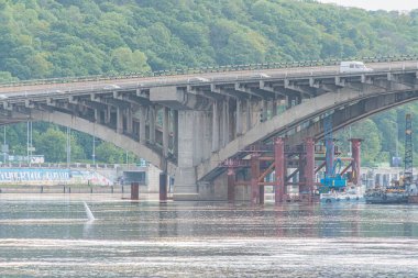 Kiev 'deki Dnepr Nehri' nin üzerinde birden fazla kemer bulunan beton bir köprünün görüntüsü. Köprü yemyeşil suların ortasında duruyor, aşağıdaki manzarayı yansıtan sakin sularla su üzerinde birkaç tekne görülüyor.