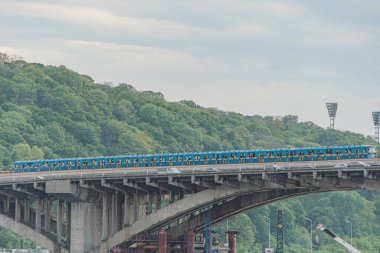 Uzun mavi tren Kiev 'deki Dnepr Nehri üzerinde beton köprüden geçiyor. Birden fazla kemer, yeşil yeşillik, ağaçlı yamaç, parçalı bulutlu gökyüzü, gerçekçilik tarzı, gün ışığı, yazı yok.