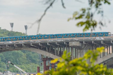 Kiev 'deki mavi Dnepr Nehri üzerinde beyaz metro köprüsü, hareket eden mavi-sarı tren şehir silueti uzaktan görülebiliyor, çeşitli boylarda ve renklerde binalar su kenarında kırmızı vinç