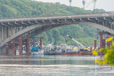 Görüntü, Kiev 'deki Dnepr Nehri üzerinde kısmen inşa edilmiş bir metro köprüsünü tasvir ediyor. Altında iki sarı vinç var. Altında dalgalar var ve bir tekne görünüyor.