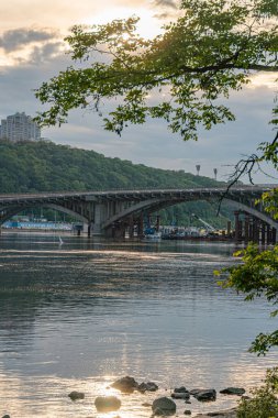 Gündoğumunda ya da batımında sakin nehir manzarası Kiev 'deki Dnepr Nehri üzerinde tarihi bir köprü sakin su, kayalık kıyı şeridi, eski köprünün birden fazla kemeri, arka planda küçük bir ada, doğal bir karışım.