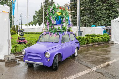 Canlı mor Volkswagen Böceği, bir etkinlik için çiçek ve kurdelelerle süslenmiş ıslak bir şehir sokağına park etmiş. Kaputun üzerinde çiçek aranjmanı, gün ışığında doğal ışıklandırmayla çekilmiş samimi bir fotoğraf.