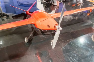 Yansıtıcı yüzeyde kırmızı kuadkopter drone, arka planda bir kişinin belirsiz figürü, kapalı alanda parlak ışıklandırma.
