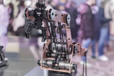 Metalik bir robotik kol, birden fazla eklemli ve bir baz tarafından genişletilen, kapalı bir ortamda sergileniyor, muhtemelen bir ticaret gösterisi veya sergisinde. Arka plan insanların bulanık figürlerini içeriyor.