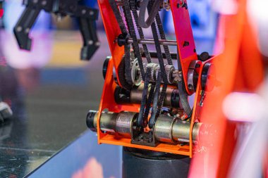 Yüzeydeki turuncu ve siyah bir robot kolunun yakından görüntüsü, karmaşık bir şekilde dişliler, kemerler, kuyular, ön plandaki insanlar, bulanık, görüntü ayarları