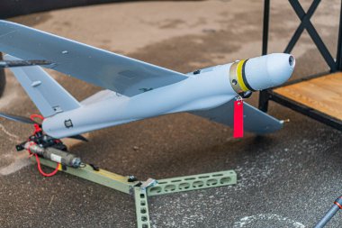 Beton sanayi hangarında asılı gri ve beyaz dron, burunda pervane, simetrik kanatlar, kuyrukta kırmızı etiket, metin yok, doküman çekimi