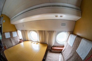 Vintage interior of a small airplane cabin with light colored walls, metal ductwork, partially drawn curtains at a window, multiple rows of white and brown seats, carpeted floor, and a central table clipart