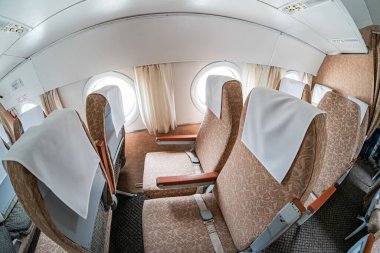 İçi boş uçak kabini, birden fazla bej kaplı koltuk, halı kaplı zemin, tepe kutuları, okuma lambaları, gövde yapısı, yapay ışıklandırma, kısmen çizilmiş pencere perdeleri, markalaşma yok.