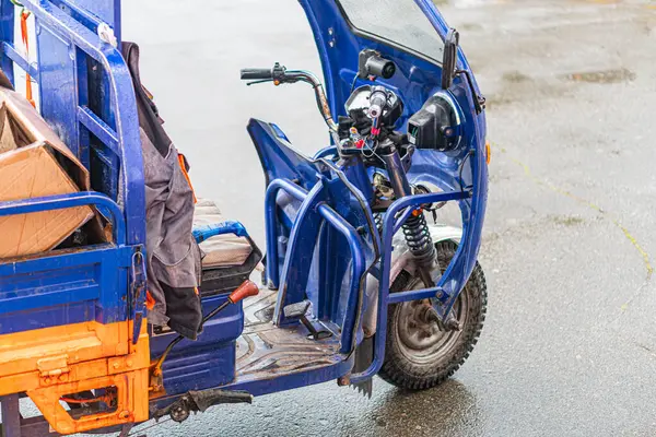 Elektrikli kargo üç tekerlekli bisiklet ve motosiklet, bulutlu gökyüzünün altında park edilmiş mavi kurye minibüsü, açık bölmeli nesneleri gösteriyor. Islak zemin, yağmurun yakın zamanda geldiğini gösteriyor.