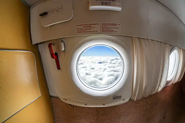Ticari bir uçağın içinden, oval bir pencereden gökyüzünü ve aşağıdaki bulutları gösteren, oturağın bir kısmı güneş ışığında görünen gizli bir görüntü.