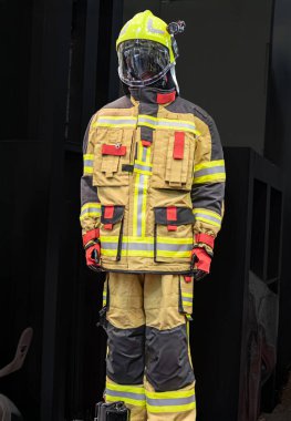 Koruyucu giysili itfaiyeci, parmaklıklı bir pencerenin önünde duruyor, gündüz ışığı, sarı kask, gözlük, yüksek görüş açısı olan ceket, sarı çizgili siyah pantolon, eldiven, alet ya da cihaz...
