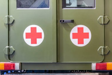 Zeytin yeşili, kırmızı haçlı askeri bir ambulansın yan görüntüsü. Kapalı bir yere park edilmiş, suni aydınlatma altında. Büyük sürgülü kapı ve faydacı tasarım elementleri görünür
