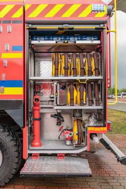 Tuğla kaldırımdaki kırmızı itfaiye aracı, sarı hortumlu ve kırmızı başlıklı açık kompartıman, metal kaplı bölmeler yerleştirilmiş ekipmanlar, dış mekan ayarı, doğal ışıklandırma, mevcut insan yok.