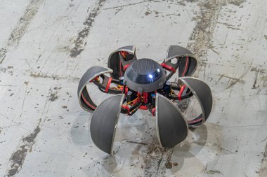 Siyah ve kırmızı robotik bir örümcek, arka ayakları üzerinde, eklemleri eklemli, kapalı beton bir ortamda, hatta ışık altında.