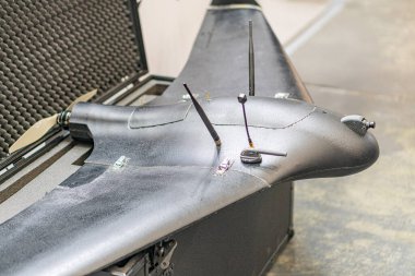 Sırtında desenli bir tabanı olan siyah bir dron. Görünür bileşenler arasında bir pil paketi, kontrol paneli ve eklentiler var. Yapay ışıklandırma bile güçlü gölgeler olmadan sahneyi aydınlatır.