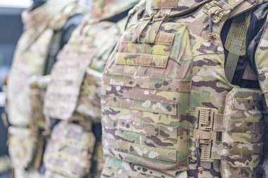 Kamuflaj kurşun geçirmez yelekli üç kişinin görüntüsü, merkezi odak noktası tek bir figür, yelekler ayarlanabilir kayışlar ve dolgulara sahip, muhtemelen askeri veya taktiksel kullanım için gerçekçi fotoğraflara sahip.