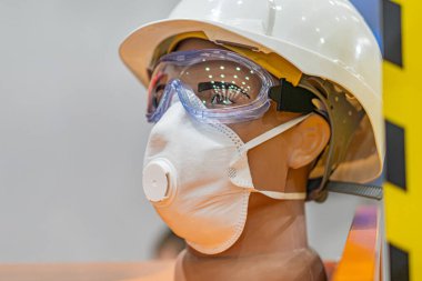 Beyaz bir miğfer, güvenlik gözlüğü ve solunum maskesi de dahil olmak üzere inşaat veya endüstriyel PPE giyen bir kişinin iç mekan ışığı ve sessiz kolo ile kapalı bir ortamda çekilmiş samimi bir yan görüntüsü.