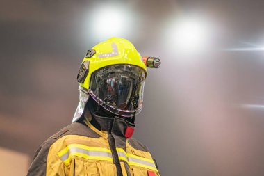 Sarı kasklı itfaiyeci, tam teçhizat, oksijen tüpü, stadyum arka planı, aydınlatılmış ışıklar, yansıtıcı çizgiler, beyaz yüz maskesi, özel birim üniforması.