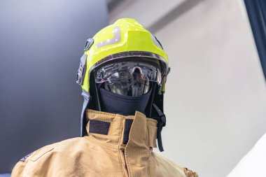 İtfaiyecilerin açık arkaplanlı, vizörlü sarı bir miğfer ve 150 numaralı bir takım elbise içinde yansıtıcı çizgili ve birden fazla cebi olan koyu renk bir ceket.