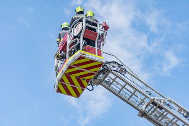 Kurtarma operasyonu devam ediyor, hava bulutlu, merdivenli itfaiye aracı, kurtarma ekibi üyeleri, miğferler, koruyucu teçhizat, kasıtlı vücut dili, yayılmış ışık, kırmızı itfaiye aracı