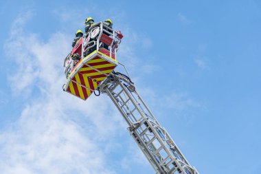 Kurtarma operasyonu devam ediyor. Geniş merdivenli beyaz itfaiye aracı, platformda iki itfaiyeci, miğferler, kırmızı ve sarı şeritler, yüksek irtifa, mesaj yok, aktif olay yerine odaklan.