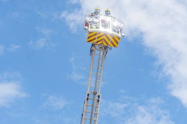 İtfaiye kurtarma operasyonu devam ediyor. Kırmızı ve beyaz çizgili merdiven kamyonu iki itfaiyeciyi kaldırıyor, kask takıyor, yansıtıcı ekipman takıyor ve emniyet kemeri takıyor.