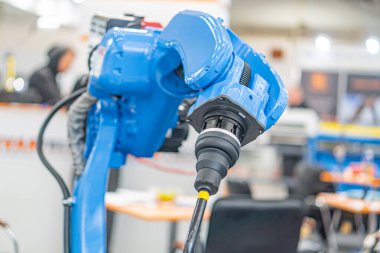 Daha çok mavi renkte bir endüstriyel robot, bir fabrika ortamında gösteriliyor. Büyük boyutlu robot, genişletilebilir bir kola ve nesne manipülasyonu için bir uç efektine sahip.