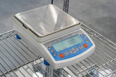 Yapay ışıklandırma altında elektronik denge ölçeği, dijital ekran, metalik kap ve diğer laboratuvar ekipmanlarının yer aldığı bir laboratuvar sahnesi.