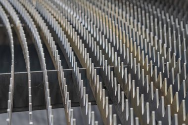 Metalik çivi ya da çivilerden oluşan kavisli bir çizginin yakın plan görüntüsü, tekrar tekrar sıralanmış ve muhtemelen işlevsel olarak sessiz renk paleti grilerin hakim olduğu, eşit ışıklandırılmış, muhtemelen yapay bir iç mekan sahnesi.