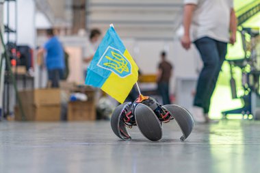 Tekerlekli robot, muhtemelen mobil, endüstriyel ayarlarda, Ukrayna bayrağı bağlı, arka planda makinelerin yanında çalışan iki kişi.