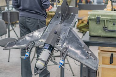 Stantta sergilenen Ukraynalı bir FPV ile kapalı mekan ayarları. Gösterişli uçak tasarımı, mat alaşımlı malzemeler, arkada küçük türbin. Koyu renk elbiseli biri.