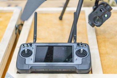 Ahşap yüzey üzerinde siyah kuadkopter drone, kontrol düğmeleri, joystickler, ekran, kapalı ortam, bulanık elektronik ekipman.