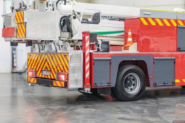 Kırmızı bir acil servis kamyonu, muhtemelen bir itfaiye kamyonu, yansıtıcı çizgili ve yangın sözcüğü görünür, kapalı bir yerde park halinde, kamyonun hidrolik vinç kolu var.