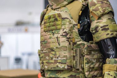 Yamalı zeytin yeşili askeri yelek. Kapalı bir kask giyen biri tarafından giyiliyor. Metal rafları ve rafları olan bir depo veya mağaza kuruyor.