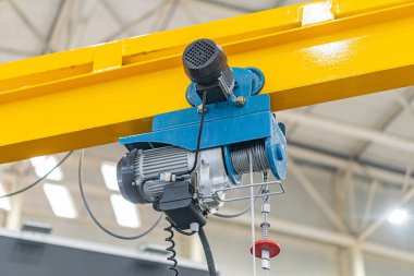 Sanayi bölgesinde çeşitli kablo ve hortumlara bağlı, üzerinde elektrikli mavi bir cihaz bulunan sarı bir vinç tasvir ediliyor. Dekor, yapay ışıklandırması olan kapalı bir fabrika.