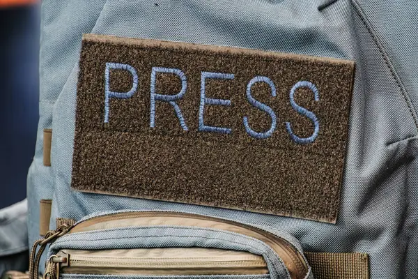 Arkadan biri, açık renk ceketli, koyu renk gömlekli ve kot pantolonlu, içinde birden fazla cebi olan zeytin yeşili bir sırt çantası taşıyan ve kapalı ya da aydınlık, açık hava ayarlı bir basın kartı olan biri.