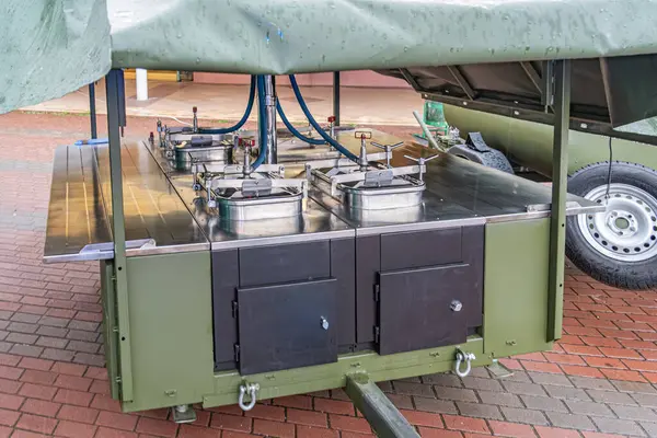 Açık havada kırmızı tuğlalı bir alanda park etmiş koyu yeşil bir yemek mutfağının samimi bir görüntüsü. Büyük bir metal tezgah, bir ızgara veya yemek pişirme istasyonu, birden fazla yakıcı ve iki set...