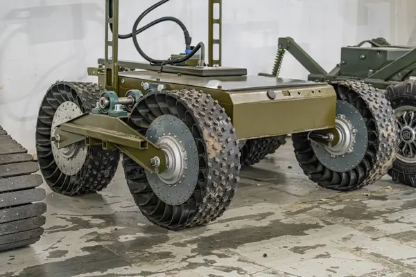 Tekerlekli, koyu renkli, beton zeminde Ukrayna askeri mini robotunun kapalı bir görüntüsü. Robotun silindirik gövdesinden çıkan kablolar ile geniş siyah izleri ve küçük tekerlekleri var. Anothe