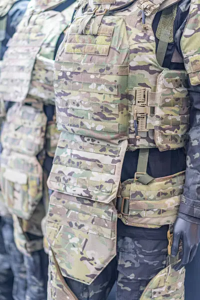 Kamuflaj yelekli, çantalı bir manken, yapay ışıklandırma altında tarafsız bir arkaplanda sergileniyor..