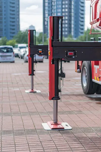 Park halindeki itfaiye aracı, indirilmiş kırmızı bariyer sistemi, cam cepheli modern mimari binalar, temiz gökyüzü, desenli tuğla kaldırım, hiç insan yok.