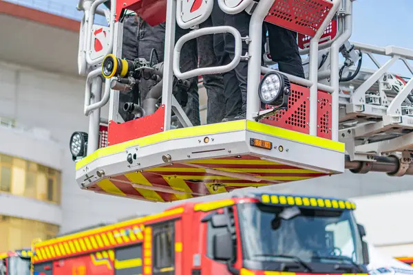 İtfaiye aracı kaldırma operasyonu, merdivendeki itfaiyeciler, basamaklı beyaz merdiven, kırmızı itfaiye aracı gövdesi, beyaz süsleme ve harf, bulutlu gün ışığı, açık hava ayarı, refleksli arka plan binası.