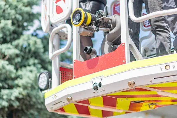 İtfaiye aracı kaldırma sahnesi, açık hava kurtarma etkinliği, merdivendeki itfaiyeci, yansıtıcı çizgili kırmızı kamyon, olası etkileşim, ağaçlar, açık mavi gökyüzü, gün ışığı.