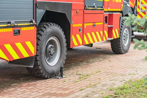 Tuğla kaldırımda kırmızı itfaiye aracı, beyaz şeritler, birden fazla kapı, açık, büyük siyah lastikler, muhtemel merdiven ya da eklenti, çevre, yeşillik, doğal aydınlatma, gündüz, yan görüş