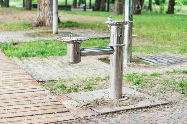 Açık havada, taş plaka üzerinde çift su fıskiyesi. Yansıtıcı, paslanmaz çelik yapı. İki sivri uçlu, sonbahar yaprakları zemini kaplıyor.