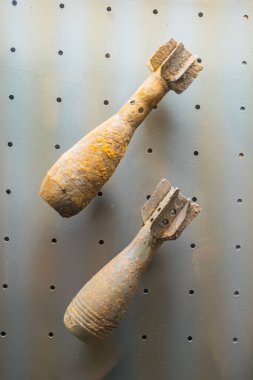 Paslı, eski topçu mermileri metalik bir yüzeye monte edilmiş. Bir kabuk önde, diğeri hafif arkada. İkisi de görünür kıvrımlı şekiller ve deliklerle donatılmış.