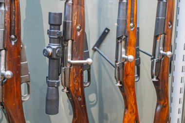 Bir müze ortamında, yatay ve eşit aydınlatılmış antika tüfekler sergileniyor. Ahşap, metal teçhizatlı yaş belirtileri gösteriyor. Yazı yok, insan yok.
