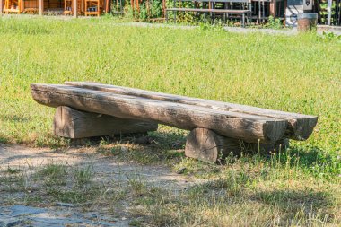 Antik Orta Avrupa köyünün dış mekan sahnesinde aşınmış ahşap bir bank üzerine odaklanması, kısmen görünür bina arkaplanı ve çimenli alana karşı. Aydınlatma şafak veya alacakaranlıkta olduğunu gösteriyor.