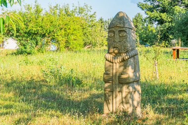 Bahçe düzeninde antik Orta Avrupa taş heykeli, biçimlendirilmiş yüz hatları ve geleneksel kıyafetleri olan antropomorfik figür, elle oyulmuş sert taş, otlar ve yabani otlar, doğal arkaplan ağaçları