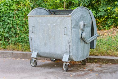 Eski Orta Avrupa köyü paslı metal atık kutusunu tekerlek ve kulplarla donatıyor, kısmen yeşillik tarafından örtülmüş çimenlik, uzaktaki ağaçlar sıcak, yumuşak li